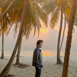 Robert Pattinson, in a stylish leather jacket, standing on a sandy beach at sunset. Surrounded by palm trees.