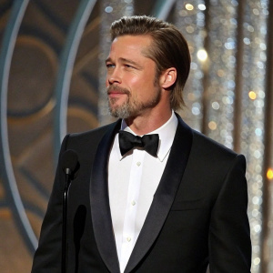 Brad Pitt, sporting a close-cropped beard and short haircut, in a sleek black tuxedo, at the Golden Globe Awards. Stands against a shimmering metallic backdrop.