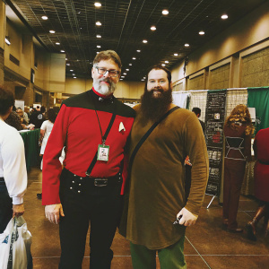 Jonathan Frakes and a bearded attendee at a Star Trek convention. The attendee wears a Starfleet uniform and sports a lanyard. The convention hall is bustling with fans and cosplayers.