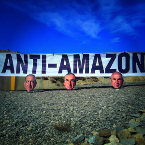 Jeff Bezos face cutouts and a large protest banner at an anti-Amazon rally. Rocky field backdrop