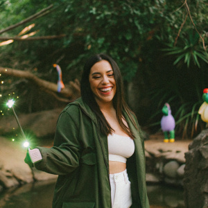 Lucy Heartfilia, from Fairy Tail, smiling and winking. Wearing a casual outfit with a green jacket and white pants. Set in a vibrant fantasy world, surrounded by magical creatures and elements.