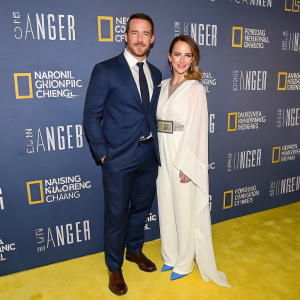Barry Sloane and Katy Sloane, posing at the National Geographic Channel event. Barry in a navy suit and tie. Katy in a flowing white top and unique belt. Blue toned shoes. Backdrop with National Geographic branding.