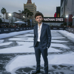 Nathan Sykes, young actor with curly hair and stubble, dressed in a tailored navy suit and crisp white shirt, stands confidently on a bustling Hollywood red carpet. Behind him, the backdrop features bold event logos and signage. Fans and press are visible in the background.