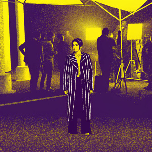 Laura Morante, a Hispanic actress, with a focused look, standing out in a bustling movie set. She wears a black and white striped jacket.