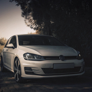 A pristine Volkswagen Golf GTI Mk 7, in a deep blue shade, parked under the dim sky surrounded by greenery, featuring its sleek aerodynamic design and the VW emblem.