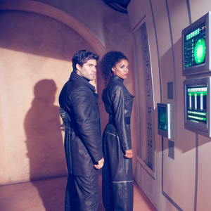Nick Zano and Maisie Richardson-Sellers in a sci-fi film scene. Dark, sleek, metallic set with futuristic technology