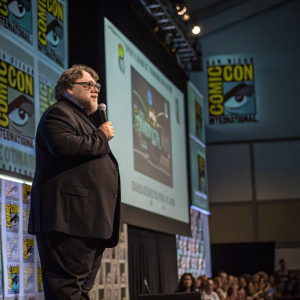 Guillermo del Toro giving a keynote speech at Comic-Con. He's dressed in a dark suit and is standing in front of a large screen showcasing various graphic elements.