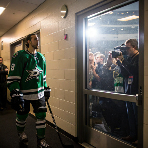 Matt Niskanen, hockey player, stands in the locker room, Dallas Stars jersey. Fans and cameras flash outside the glass window.