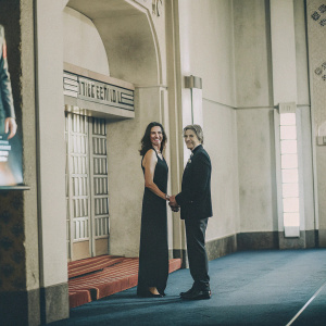 Terry Farrell and Adam Nimoy at a Star Trek themed gala. Terry wears a black floor-length dress. Adam in a dark suit. Blue carpet and Star Trek logos in the background. They pose for a formal photo.