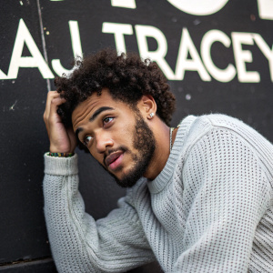 Close-up shot of Aj Tracey. African male with curly hair and beard. Playful pose. Gray knit sweater. Black wall with white text backdrop