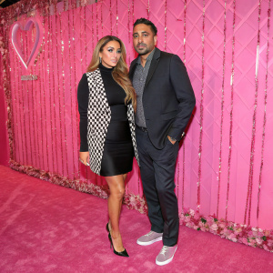 Larsa Pippen and Umar Kamani at a fashion gala. The backdrop is a vivid pink wallpaper with shimmering details. Larsa wears a black dress with a patterned vest, standing confidently.