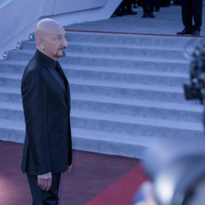 Sir Ben Kingsley, bald head, sharp jawline, and neatly groomed facial hair. Dressed in a sleek black suit. Attending a prestigious film festival.
