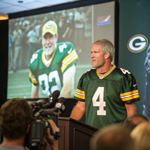 Brett Favre in a press conference. Wearing a green jersey with number 4. Behind him is a large screen displaying highlights of his career.