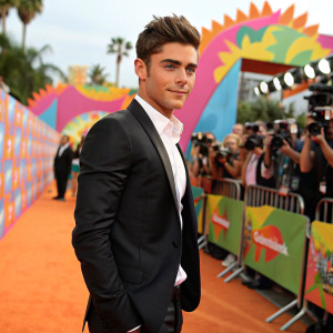Zac Efron, dressed in a sleek black suit with a crisp white shirt, posing for photographers at a vibrant outdoor Nickelodeon event.