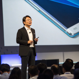 Koh Dong Jin, Samsung executive, addressing a tech conference. Black suit, white shirt. Large screen behind, showcasing Galaxy Note 7