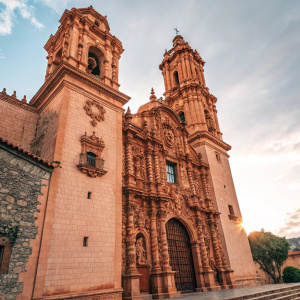 Photograph of the Santa Prisca Church in Taxco, Guerrero, Mexico. Ornate architecture with intricate carvings in warm terracotta hues. Central ogee archway frames detailed window sculptures. Rich textures and elaborate designs highlight the building's facade.
