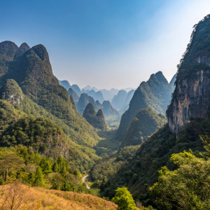 A panoramic view of the Ha Giang karst geopark. Towering, jagged mountains with dense forests. Clear blue sky, lush vegetation in shades of green and brown covering the steep slopes.