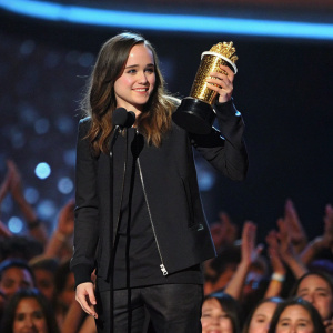 Ellen Page, standing on stage at MTV Movie Awards. Holding golden trophy, dressed in sleek black jacket. Audience cheering in background.