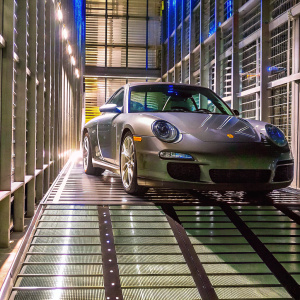 A glossy, modern gray Porsche GT3 RS parked in an upscale garage. The sleek car stands out against the structured grid-like floor, surrounded by a robust metal framework and diffused lighting, casting dramatic shadows.
