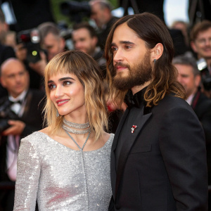 Jared Leto and Sofia Boutella at Cannes Film Festival premiere. Jared in a black tailored suit, Sofia in a sparkling silver dress. Crowded venue with flashing cameras