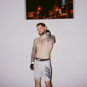 Renowned MMA fighter Frankie Edgar, sporting a rugged beard and multiple tattoos. Attired in Reebok UFC shorts. Stands confidently in a serious pose at a promotional event. Backdrop: neon-lit cityscape