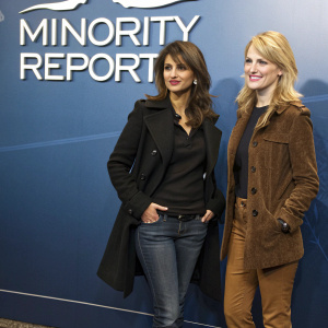 Monica Cruz and Kathryn Morris at a promotional event for 'Minority Report' in Madrid, Spain. Monica in black coat and jeans with a serious expression. Kathryn in brown jacket and pants looking cheerful. They stand in front of a blue backdrop featuring 'Minority Report'.