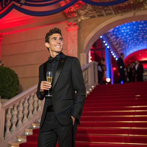 Marc Marquez at the Monaco Grand Prix after-party, dressed in a sleek black tuxedo. Champagne in hand. Lavish setting with red and blue decor.