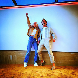 Brittany Snow and Zac Efron at the Netflix premiere. Wearing casual outfits. Behind them, a plain white wall with neon signs. Image captures their excitement.