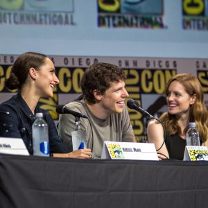 Gal Gadot, Jesse Eisenberg, and Amy Adams sitting at a table with water bottles and name cards at Comic-Con. Comic-Con logos and banners in the background.
