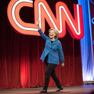Hillary Clinton waving enthusiastically on stage, large CNN logo behind her. Red stage curtains and vibrant background