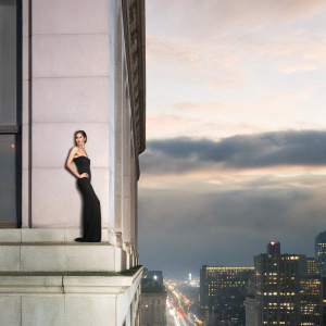 Maia Mitchell, an actress, poses elegantly. Wearing a form-fitting black dress. The background is a bustling cityscape at night.