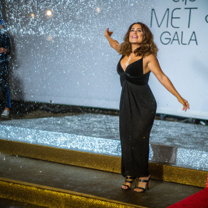 Salma Hayek in a stunning black dress with strappy sandals. Posing confidently at the Met Gala. Red carpet and glitzy backdrop featuring logos and text.