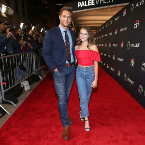 Justin Hartley and Isabella Justice Hartley, walking on the vibrant red carpet of the PaleyFest gala. Justin, in a navy blazer, paired with a patterned tie and simple brown shoes, stands beside Isabella, wearing a striking red off-the-shoulder top and sky blue jeans.