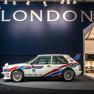 Vintage Lancia Delta S4 Rally car from 1985. On display at classic car exhibition. Dark blue backdrop, with large 'LONDON' letters in bold. Indoors, lights shining on car