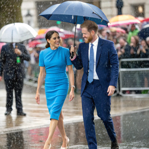 Meghan Markle and Prince Harry walking in rain. Meghan in blue dress, Prince Harry in navy suit. Crowds cheering