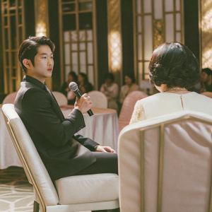Taec-Yeon, Korean actor, in a formal interview. Wears a dark suit. Event hall with gold accents and audience