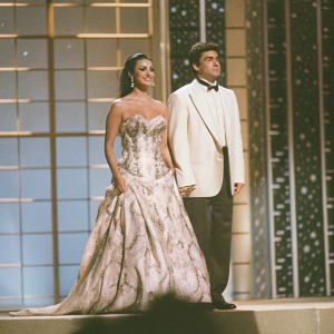 Maria Grazia Cucinotta and Alberto Tomba at the World Music Awards, Monaco, 1996. Blue gradient backdrop with golden accents. Maria in a strapless gown adorned with floral patterns. Alberto in a classic tuxedo. Both standing on a stage.