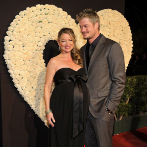 Rebecca Gayheart and Eric Dane at the annual Hollywood Valentine's Day Gala. Rebecca, pregnant, in a strapless black gown adorned with a bow, Eric in a tailored suit. They stand before a heart-shaped backdrop made of white roses. The event is illuminated by soft, ambient lighting.