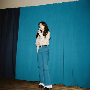 Jessica Jung, Korean-American artist, in a casual outfit, standing on stage. Talking into a microphone. Backdrop is a solid black curtain.