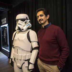 Hispanic man, mustache, maroon sweater, standing with a Stormtrooper costume. Dark background with curtain details.