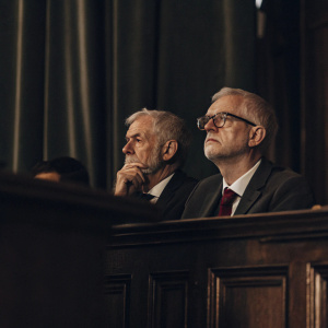 Formal photo of Jeremy Corbyn and John McDonnell at Labour Party conference. They wear suits, stand against green backdrop, serious expressions.