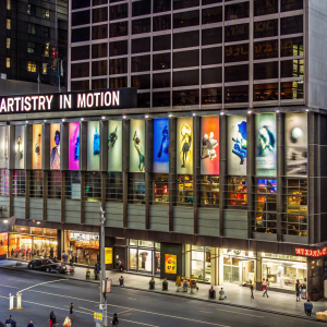 An exterior view of a dance studio called Artistry in Motion. Vibrant banners and dance-themed window decals. Located in a bustling commercial zone with ample parking.