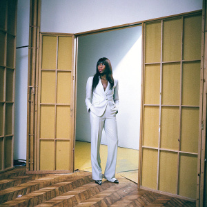 Naomi Campbell, standing solo, wearing a chic white suit. The setting is a minimalist white room.