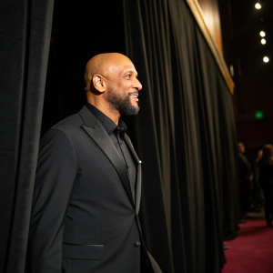 Roberto Carlos, a man with a bald head and beard, wearing a black suit. At a gala event, standing alone in front of a black curtain.