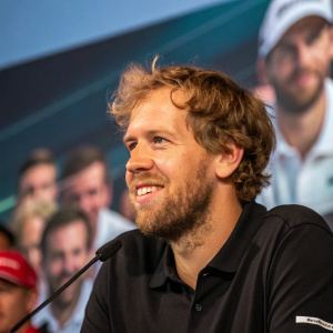 Sebastian Vettel, bearded, friendly expression, wearing a black polo shirt. Press conference, blurred audience.