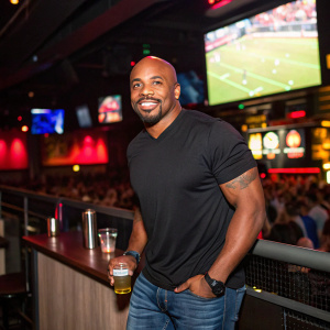 Kevin Faulk, ex-NFL player, in a casual black t-shirt and jeans. Partying at a sports bar, vibrant crowd in the background.