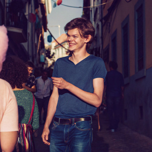 Thomas Brodie-Sangster, smiling at a bustling street festival. Wearing a blue t-shirt and jeans. Crowded urban backdrop