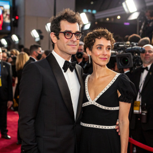 Adam Mosseri and Monica Mosseri at a Hollywood gala. Adam, in a sharp tuxedo, Monica, in a black dress with white details. The scene is vibrant with flashing cameras and celebrities.