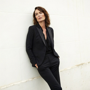 Lena Headey, standing confidently against a plain white wall, dressed in a sophisticated black suit. She wears minimal makeup, her brown hair styled loosely over her shoulders. This close-up portrait captures her intensity and charisma, perfect for a filmography page.