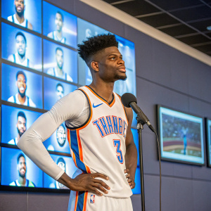 Nerlens Noel during a press conference, dressed in his Oklahoma City Thunder jersey. Behind him is a wall of screens displaying team highlights.